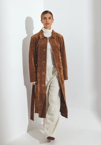 Cappotto lungo in suede marrone con colletto, chiusura a bottoni e tasche laterali, abbinato a pantaloni leggeri in lino e un maglione a collo alto bianco.