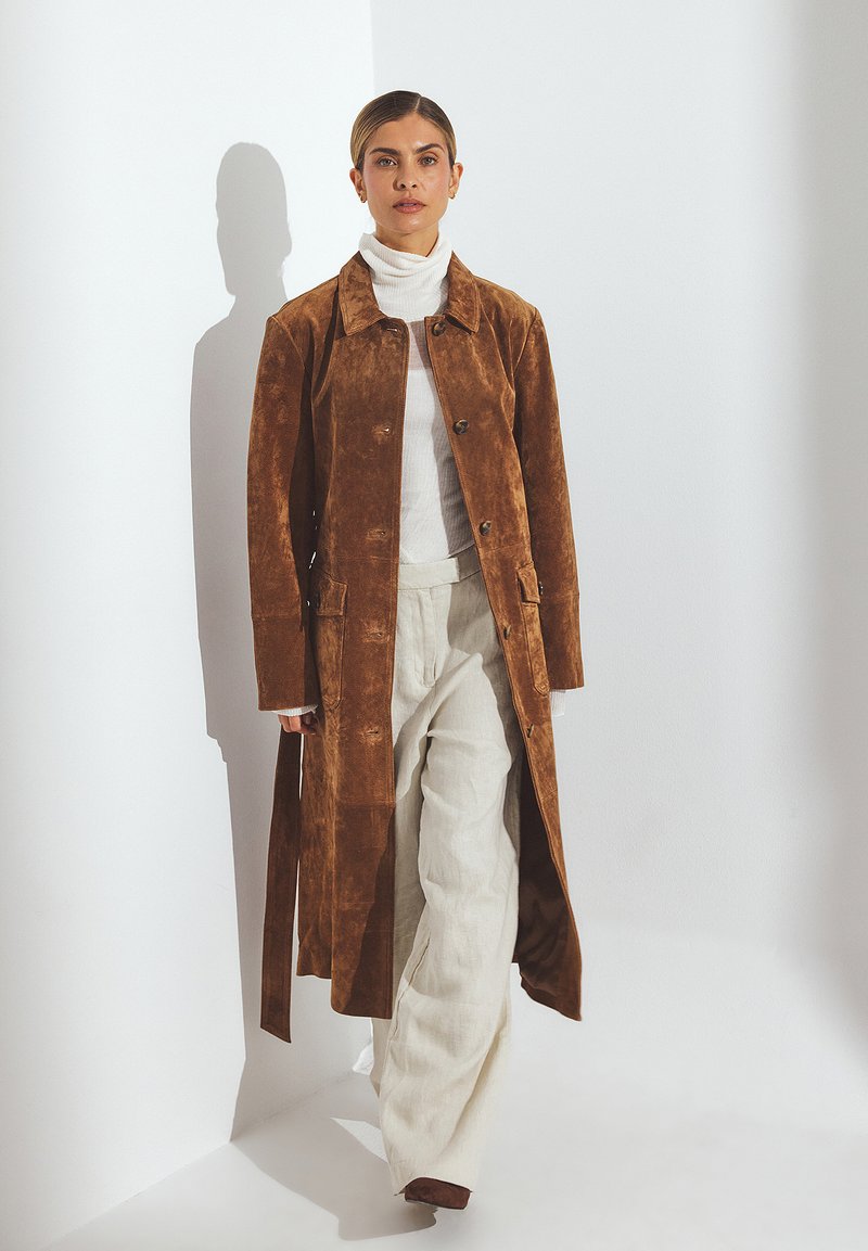 Cappotto lungo in suede marrone con colletto, chiusura a bottoni e tasche laterali, abbinato a pantaloni leggeri in lino e un maglione a collo alto bianco.