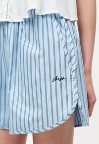 Ανοιχτό μπλε σορτς με κάθετες σκοτεινές ρίγες, πλαϊνές τσέπες και μαύρο κεντημένο αυθεντικό λογότυπο στον αριστερό μηρό, φορεμένο με λευκή μπλούζα.