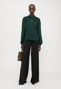 Dark green blouse with puffed sleeves and keyhole neckline detail, paired with loose black pants and a small olive green handbag.