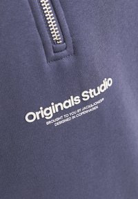 Námornícky modrý textil so zipsom a textom "Originals Studio" v bielej farbe. Hladká textúra a minimalistický dizajn.