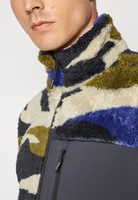Fleecejacke mit einem Camouflage-Muster in Beige, Grün, Schwarz und Blau. Hoher Kragen mit Reißverschluss und seitlicher Reißverschlusstasche in einer einfarbigen dunklen Farbe.