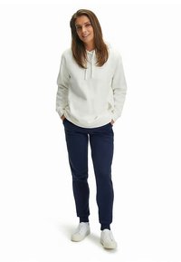 Witte hoodie met een zak aan de voorkant en een capuchon met trekkoord, gecombineerd met navyblauwe broek en witte sneakers. Geplaatst tegen een effen witte achtergrond.