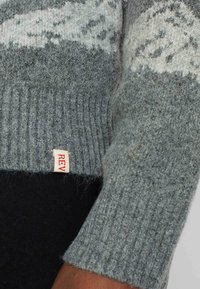 Grauer Wollpullover mit einem gerippten Bund, der ein leichtes Muster aufweist und ein kleines rotes "REV"-Etikett in der Nähe des Saums hat. Weicher Stoff, lässiges Design.