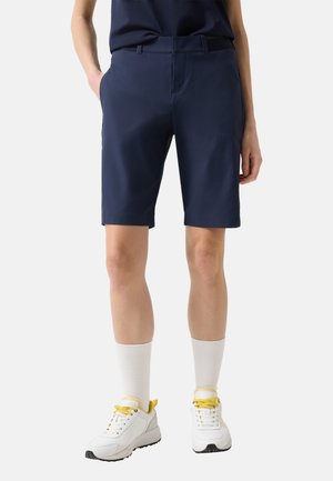 Persoon met donkerblauwe knielange shorts, witte sokken halverwege het onderbeen en witte sneakers met gele veters en accenten.