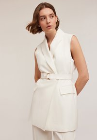 Blazer alb fără mâneci cu un design înfășurat, cu un brâu la talie prevăzut cu un inel auriu, având două buzunare aplicate și o textură de țesătură netedă.