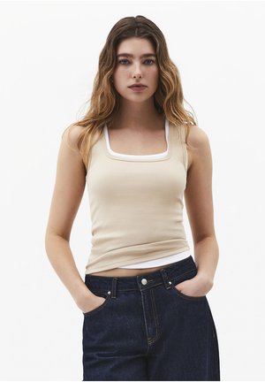 Jeune femme aux cheveux ondulés porte un débardeur beige côtelé à col carré par-dessus un sous-vêtement blanc et un jean bleu foncé, les mains dans les poches.