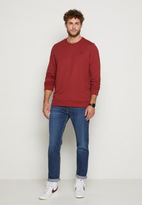 Maroone Sweatshirt mit Rundhalsausschnitt, gerippten Bündchen und Saum. Kombiniert mit blauen Jeans und weißen Sneakers mit einem maroonen Akzent.