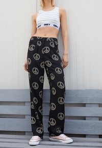 Schwarze Hose mit einem Muster aus Peace-Zeichen, weit geschnitten und aus leichtem Material. Kombiniert mit einem weißen, gestreiften Crop-Top und rosa Turnschuhen.