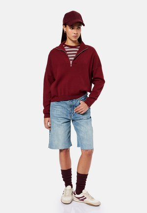 Vrouw draagt een kastanjebruine pet, kastanjebruine trui met rits, gestreept overhemd, spijkerbroek korte broek tot op de knie, kastanjebruine sokken en witte sneakers, staand met hand in zak.