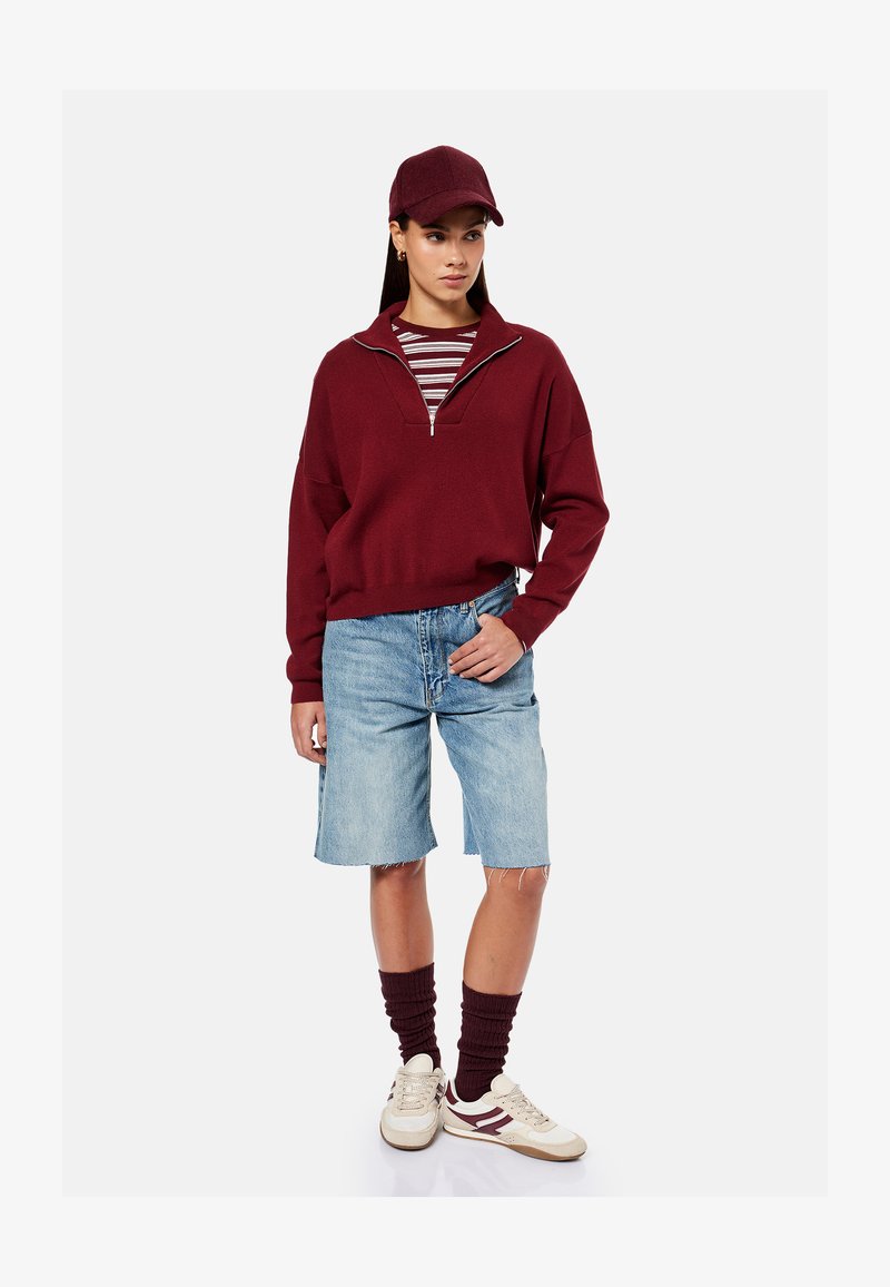 Femme portant une casquette bordeaux, un pull zippé bordeaux, une chemise rayée, un short en jean jusqu'aux genoux, des chaussettes bordeaux et des baskets blanches, debout avec la main dans la poche.