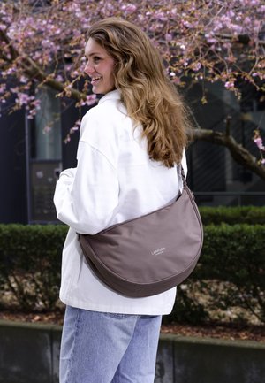 Femeie zâmbitoare cu păr lung, purtând o jachetă albă și blugi deschiși la culoare, poartă o geantă de umăr taupe Larkson mare în aer liber, aproape de un copac înflorit.