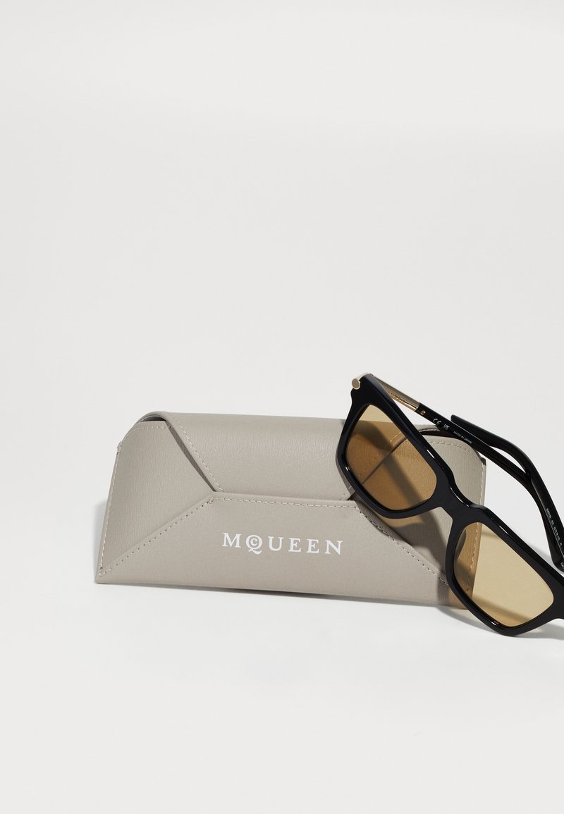 Schwarze rechteckige Sonnenbrille mit bernsteinfarbenen Gläsern, die auf einem beigen Lederetui mit McQueen-Logo auf einer weißen Oberfläche liegt.
