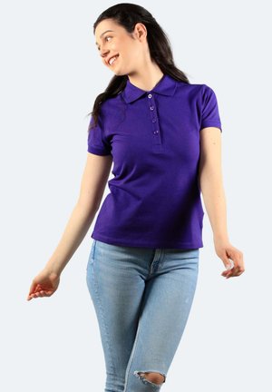 Junge Frau lächelt, trägt ein violettes Polo-Shirt mit kurzen Ärmeln und zerrissene hellblaue Jeans, steht mit leicht ausgestreckten Armen.