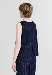 Une personne avec des cheveux châtains clairs courts porte un haut sans manches bleu marine à rayures et un pantalon assorti, de dos contre un fond simple.