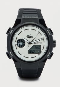 Μαύρο ψηφιακό ρολόι με στρογγυλό καντράν και λευκές λεπτομέρειες. Διαθέτει υφασμένο μαύρο λουράκι, λογότυπο και πολλαπλά κουμπιά λειτουργιών. Η οθόνη δείχνει την ώρα.