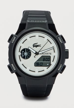 Musta digitaalinen kello pyöreällä kellotaululla ja valkoisilla yksityiskohdilla. Kellossa on teksturoitu musta ranneke, logo ja useita toimintonäppäimiä. Näytöllä näkyy aika.