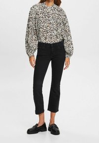 Mujer con blusa blanca de manga larga con estampado animal en negro y marrón, vaqueros negros cortos y mocasines negros, posando sobre un fondo liso.