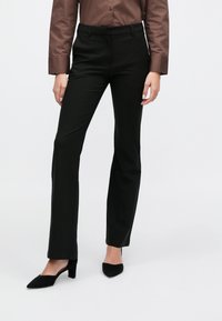 Pantalon noir sur mesure avec une texture lisse, présentant une taille ajustée et une coupe droite, accompagné de talons noirs à bout pointu.