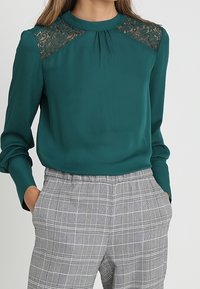 Tealblus med hög krage, spetsdetaljer på axlarna och långa ärmar, kombinerad med grå rutiga byxor med avslappnad passform.