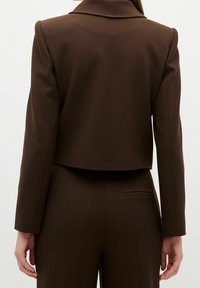 Brun kort blazer med spetskrage, långa ärmar och slät textur. Har en enda ryggslits och skräddarsydd passform.