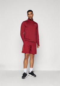 Camiseta deportiva roja de manga larga con media cremallera, combinada con pantalones cortos rojos a juego que presentan un logo, llevada con zapatillas negras y calcetines blancos.