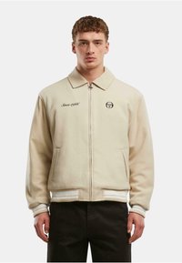 Šviesiai smėlio spalvos užtrauktukas su apykakle, šoninėmis kišenėmis, nertomis rankovėmis ir apačia, su tekstu "Nuo 1966 metų" ir logotipu ant krūtinės.