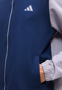 Άτομο φοράει μια ναυτική μπλε υφασμάτινη ζακέτα με λευκό φερμουάρ και λογότυπο, και ένα ανοιχτό γκρι μακρυμάνικο πουκάμισο, χέρι στην τσέπη της ζακέτας.