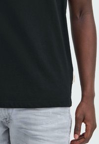 Svart bomull t-skjorte med rett snitt, som har en lett kant og en liten vevd etikett nederst. Lyse grå slitte jeans er synlige.