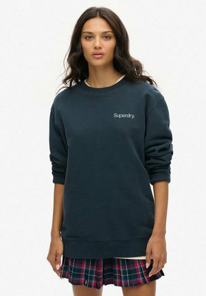 Joven con cabello largo y oscuro que lleva una sudadera Superdry azul oscura y una falda de cuadros rojos y azules, de pie contra un fondo blanco.