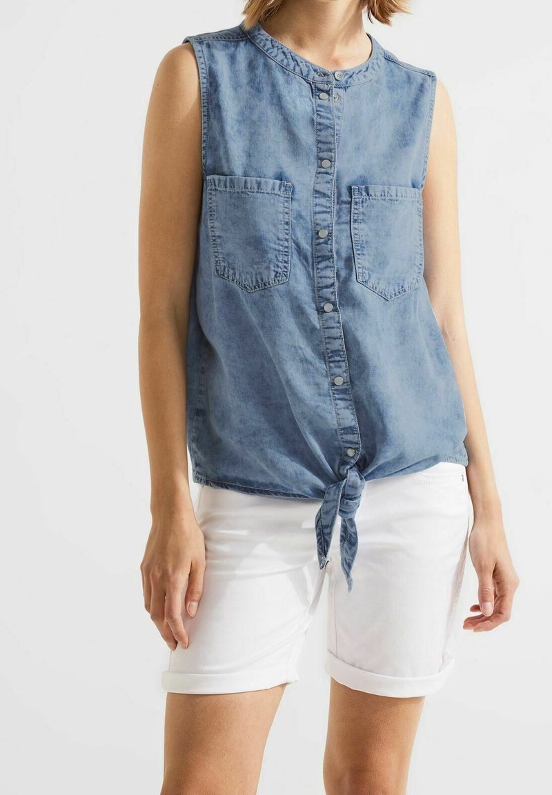 Vaaleansininen farkkaliivi, jossa on napit edessä, kaksi rintataskua ja solmittu vyötärö, yhdistettynä käärittyihin valkoisiin shortseihin.