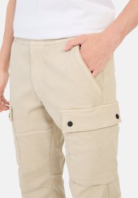 Beige Cargo-Hosen aus Baumwolle, mit seitlichen Taschen und schwarzen Druckknöpfen, sichtbaren Nähten und einem elastischen Bund für Komfort.