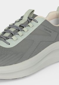 Gros plan d’une basket Calvin Klein grise et vert clair avec une semelle arrondie et des lacets bouclés sur fond clair.