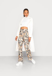 Calças flare estampadas em tons de bege, castanho e azul com padrões em espiral; combinadas com um suéter cropped branco e um casaco longo branco. Ténis brancos completam o visual.