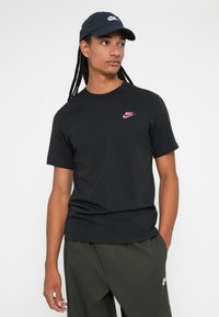 Juodas medvilninis marškinėliai trumpomis rankovėmis su mažuoju rožiniu Nike logotipu ant krūtinės. Dėvimi kartu su alyvuogių spalvos kelnėmis ir atitinkančiu kepuraitę.