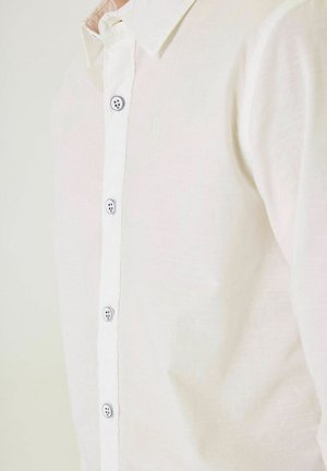 Chemise boutonnée beige clair en tissu texturé, dotée d'un col classique, d'une patte de boutonnage blanche et de boutons sombres sur le devant.