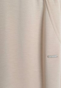 Beiger Stoff mit glatter Textur und dezentem Muster. Verfügt über eine Seitentasche und ein kleines silbernes Logo-Label in der unteren rechten Ecke.