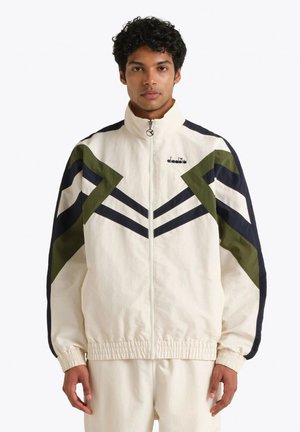 Uomo che indossa una giacca bianca con cerniera e strisce a chevron verdi e blu navy, abbinata a pantaloni bianchi, in piedi davanti a uno sfondo neutro.