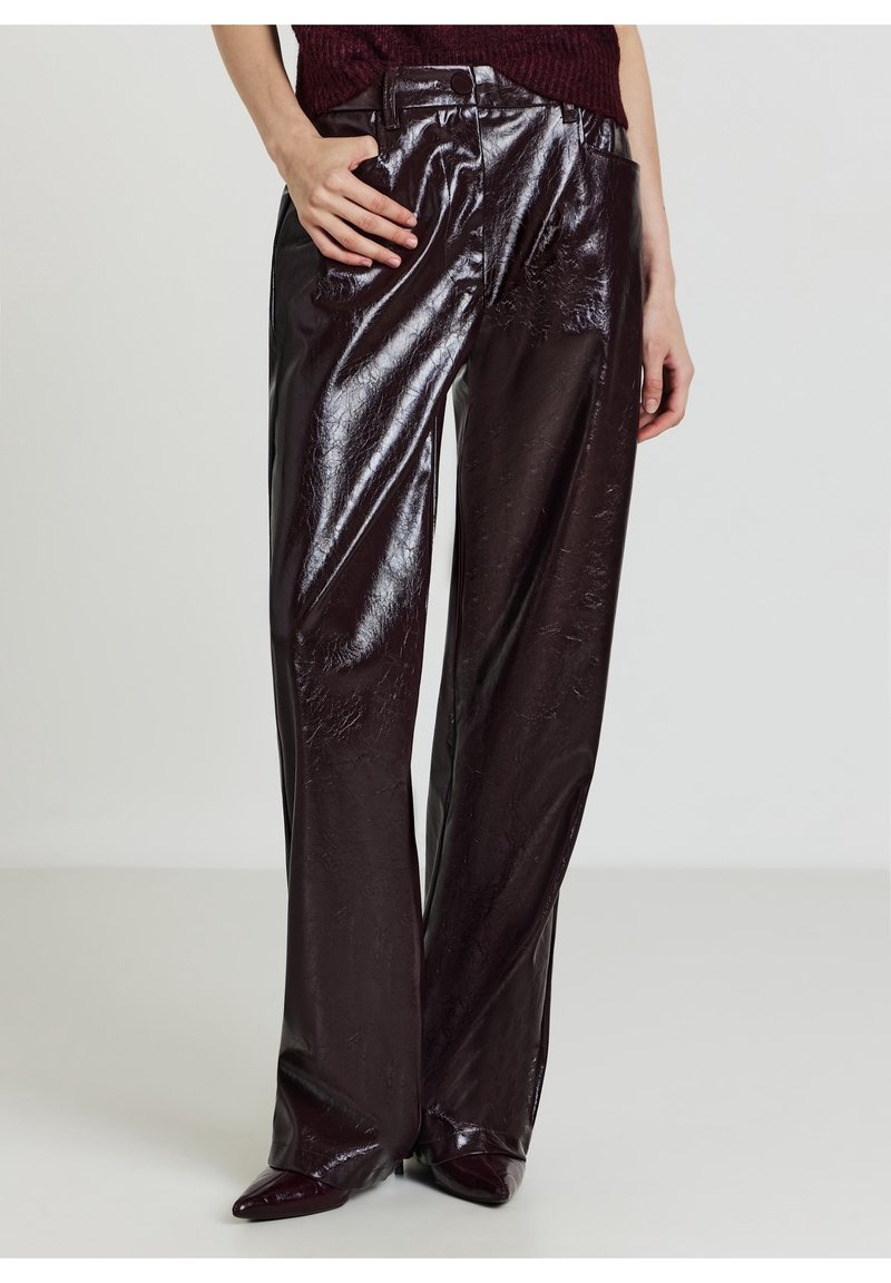 Pantalon large marron foncé brillant avec une texture froissée, porté avec des chaussures pointues et un haut bordeaux rentré, main du modèle sur la hanche.