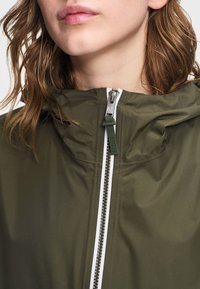 Κοντινό πλάνο ενός ατόμου που φοράει ένα μπουφάν με κουκούλα σε ελιά πράσινο χρώμα, με λευκή επένδυση στο μπροστινό φερμουάρ και χαλαρά κυματιστά μαλλιά.