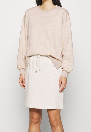 Mujer con una sudadera beige de manga larga y hombros caídos, y una falda crema de longitud hasta la rodilla con cordón ajustable, posando frente a un fondo liso.