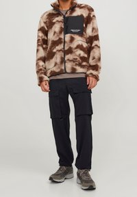 Brun och beige fleecejacka med dragkedja och bröstficka. Buren med svarta cargobyxor och grå sportskor.