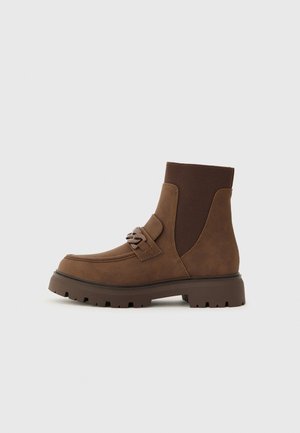 Bottines marron avec un dessus lisse et texturé, des panneaux latéraux élastiques et une semelle épaisse à crampons. Présente un détail décoratif de sangle.