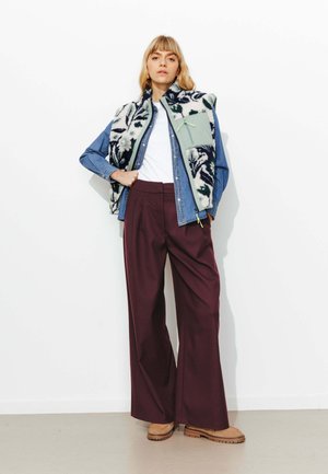 Chemise en denim superposée sous un gilet en polaire à motif avec une poche verte, associée à un pantalon large bordeaux et des chaussures à enfiler couleur tan.