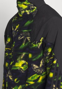 Giacca in pile con tessuto nero e un vivace motivo floreale verde e giallo. Presenta un colletto alto e strisce laterali. Chiusura frontale con zip.