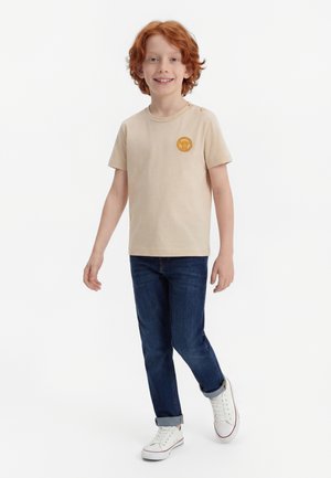 Smilende rødhåret gutt som går, iført beige t-skjorte med gul lapp, oppbrett mørkeblå jeans og hvite joggesko mot en ensfarget bakgrunn.