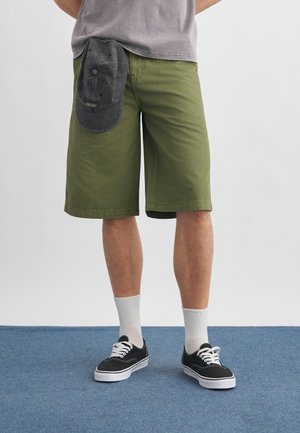 Persona que lleva pantalones cortos verde oliva, zapatillas negras con cordones blancos, calcetines blancos y una gorra gris colgando del bolsillo de los pantalones cortos.