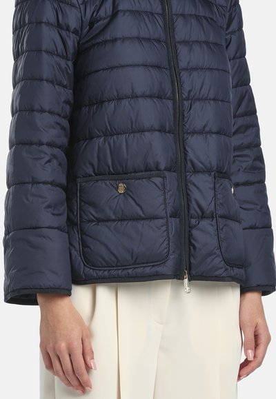 Donna che indossa una giacca trapuntata blu navy con zip frontale e tasche applicate con bottoni, abbinata a pantaloni beige chiaro.