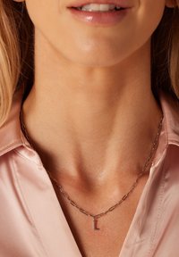 Zilveren ketting met verlengde schakels en een klein letter "L" hangertje. Draagt over een lichtroze satijnen blouse.