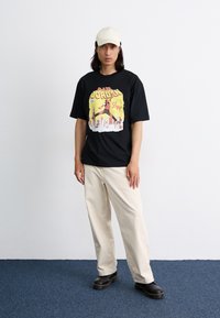 T-shirt noire avec une impression graphique colorée d'une figure en train de sauter et du texte. Associée à un pantalon ample de couleur crème et des chaussures noires.
